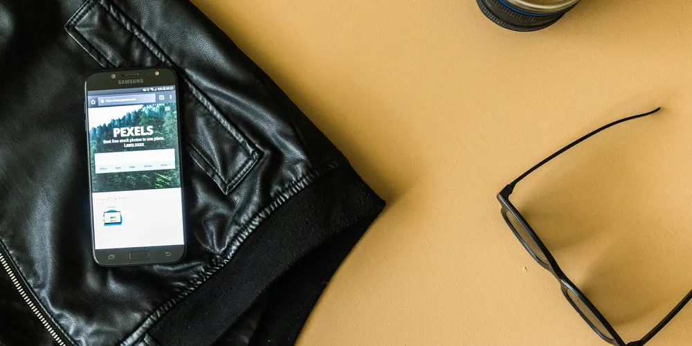 Flat lay of a black leather jacket with a smartphone showing a forest image and a pair of sunglasses on a tan surface.