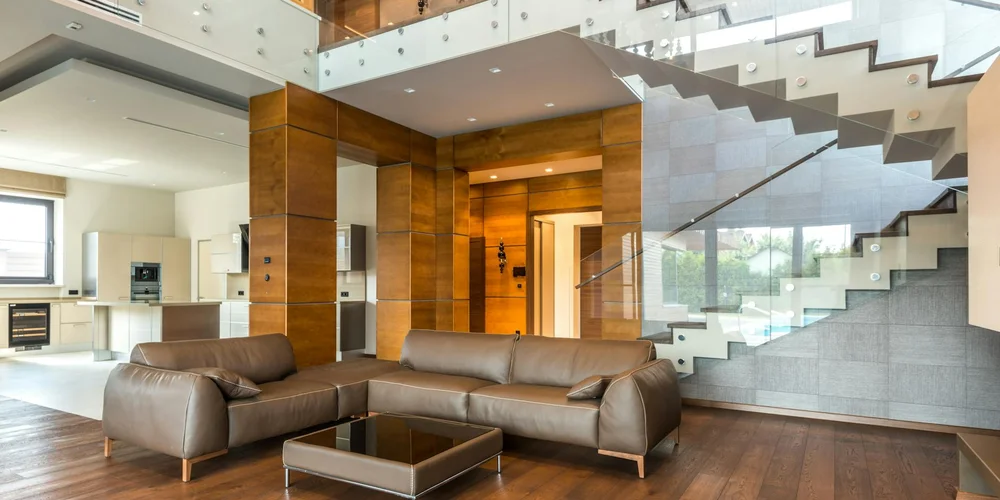 Modern living room with a brown leather sectional sofa, glass coffee table, and a staircase with glass and metal railings.