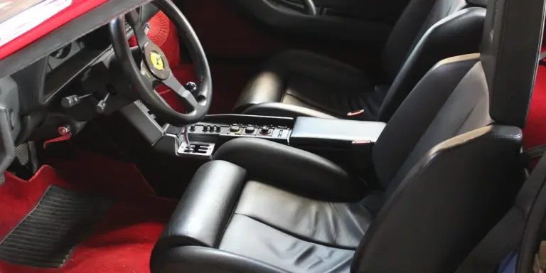 Close-up of a luxury car interior: black leather seats, a steering wheel with a Ferrari logo, and red floor carpeting.