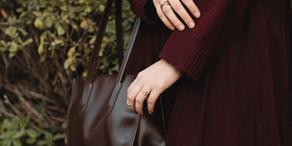Close-up of a burgundy coat with a brown leather handbag, with hands resting on the bag to evoke careful leather maintenance.