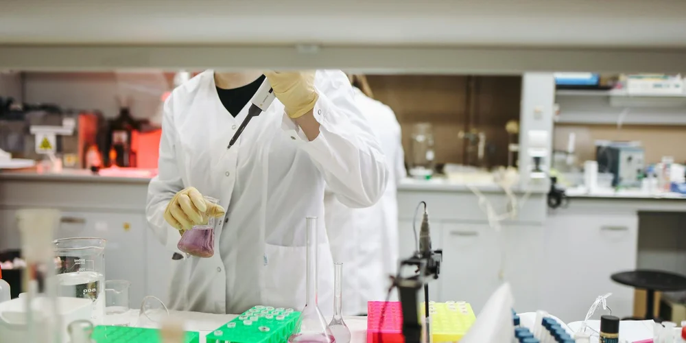 Lab technician in a white coat and gloves handling a purple liquid in a beaker, with test tubes and lab equipment in the background.