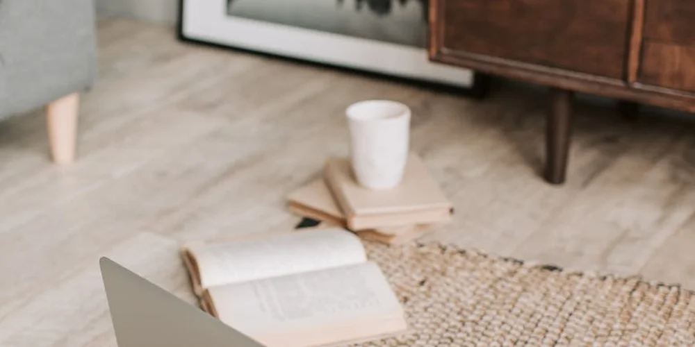 A light-colored living room with a woven jute rug, an open book, a white mug, and a small stack of books on pale wood flooring.