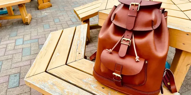 Brown leather backpack resting on a wooden outdoor bench.