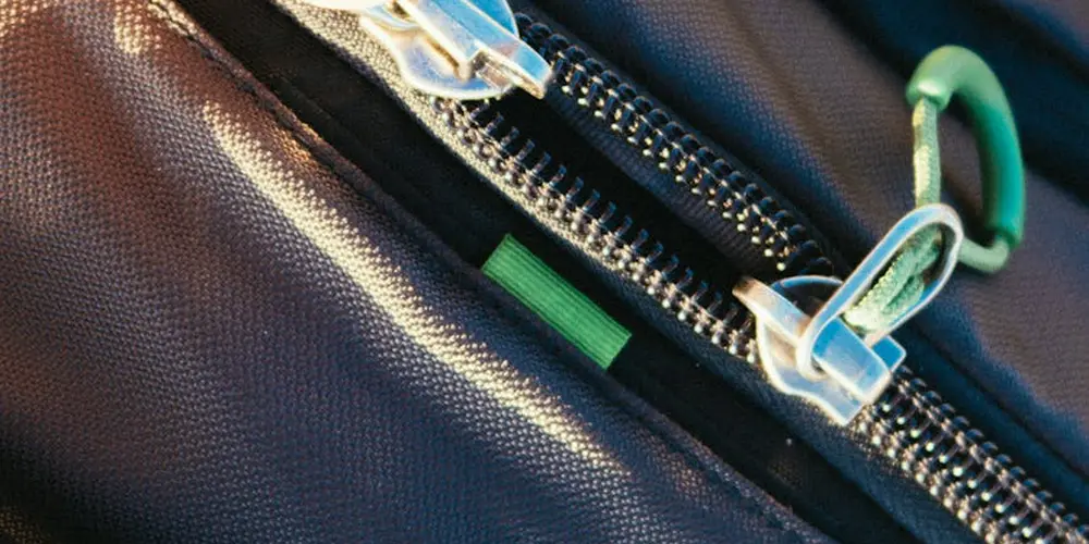 Close-up of a leather bag featuring metallic zippers, stitching, and a green pull tab, highlighting premium materials.