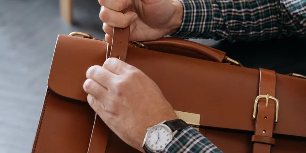 Close-up of hands adjusting the strap on a brown leather bag, with a plaid shirt visible in the background.