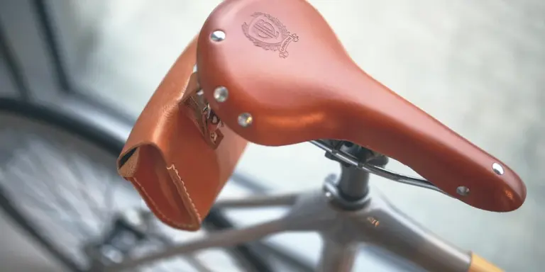 Close-up of a brown leather bicycle saddle with a small attached bag, showing leather grain and rivets.