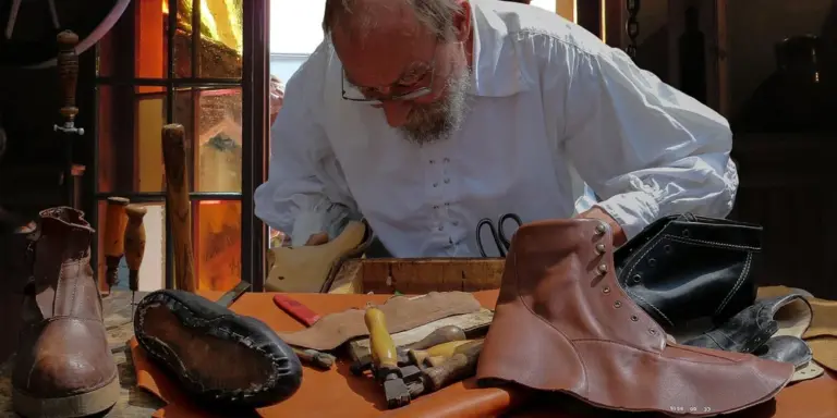 A craftsman with a white shirt works on leather boots at a cluttered workbench, surrounded by tools, in a workshop.