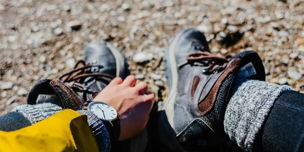 Close-up of grey leather boots on rocky ground with a person wearing jeans and a yellow sleeve, suggesting preparation for cleaning.