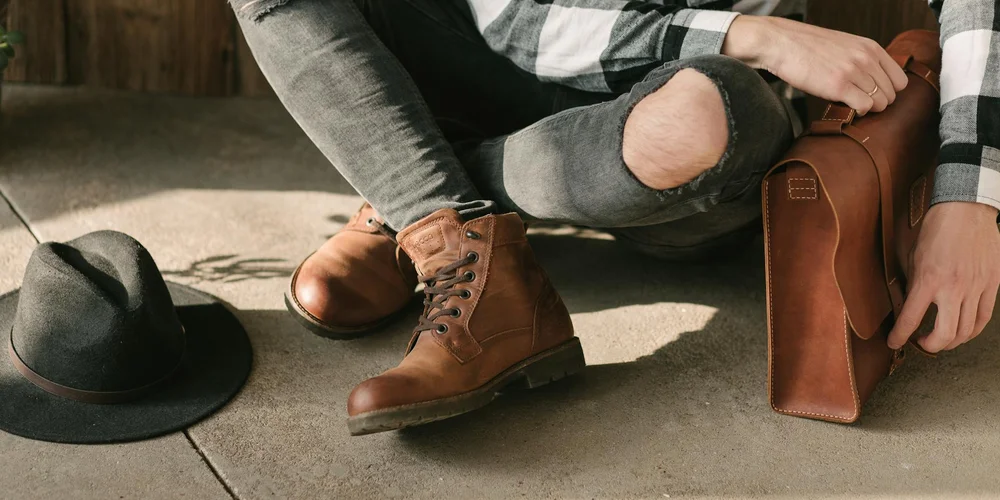 Brown leather boots resting on a concrete surface next to a bag; a person sits nearby wearing jeans and a plaid shirt.