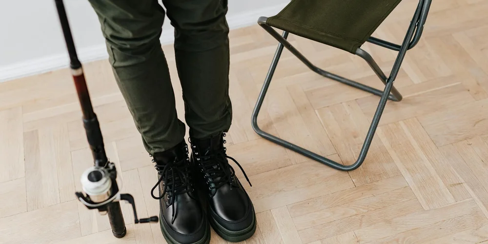 Close-up of black leather boots with laces on a light wood floor; a folding chair and a fishing rod are visible in the background.