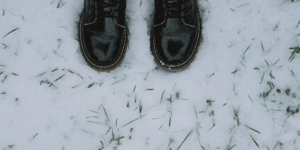 Top-down view of black leather boots standing in light snow with a few blades of grass visible around them.