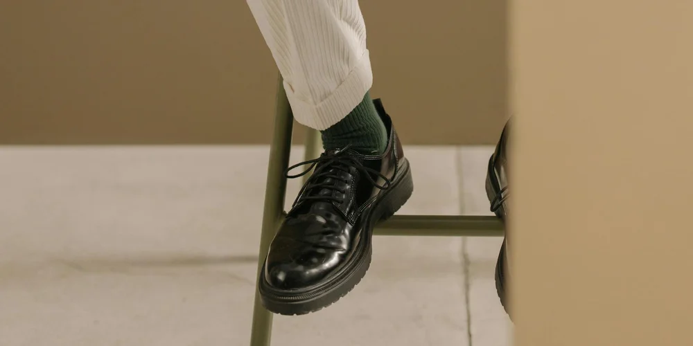 Close-up of black leather dress shoes resting on a chair, with a person wearing light-colored trousers; cardboard boxes in the background.