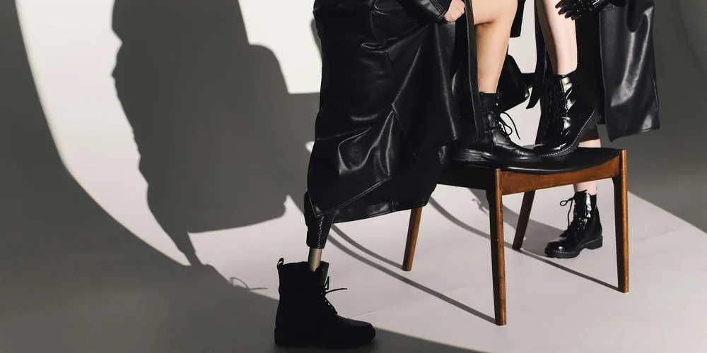 Black leather boots and jackets arranged in a fashion shoot with a wooden chair, dramatic shadows, and a gray studio backdrop.