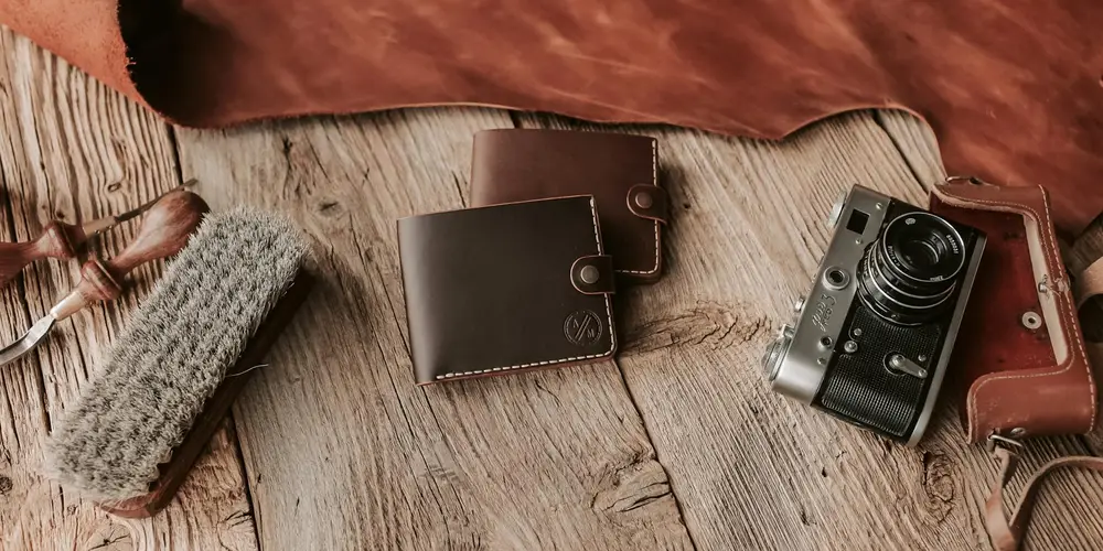 Wallet, belt, and other leather items laid on a wooden surface with a leather brush and a vintage camera nearby