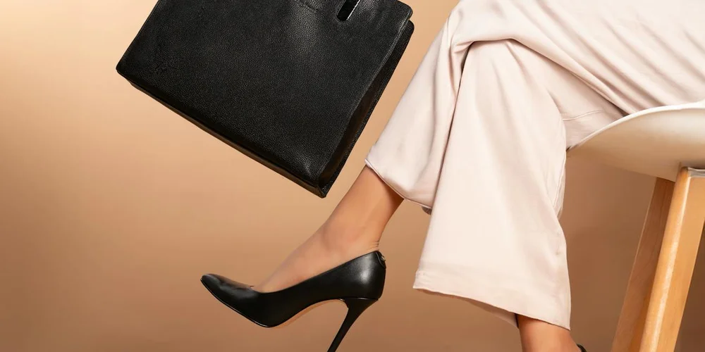 A person wearing beige pants and black high-heeled shoes sits beside a black leather bag, highlighting leather footwear and accessories.
