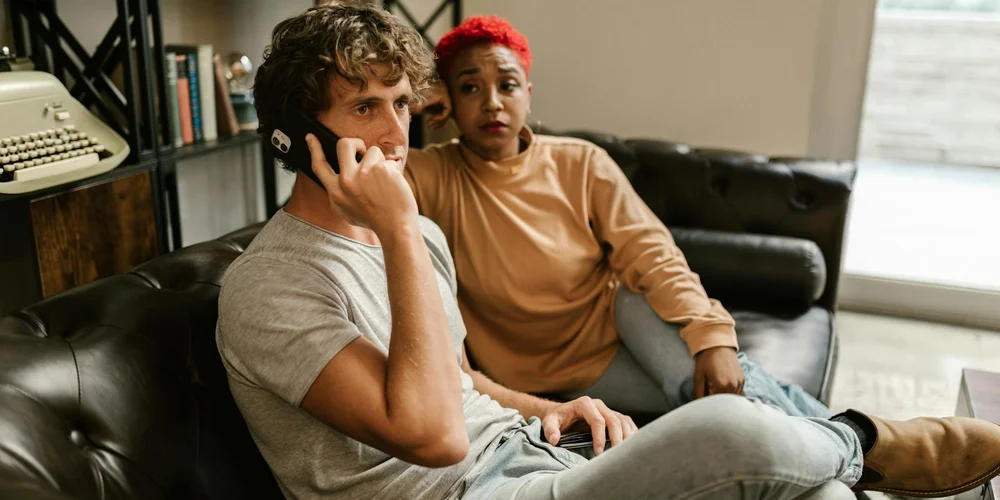 A couple sits on a black leather sofa; one person is on the phone and the other looks concerned, suggesting a potential leather couch repair issue.