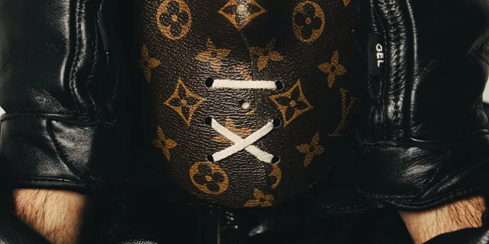 Close-up of black leather gloves worn by hands, with a Louis Vuitton monogram-pattern item in the background.