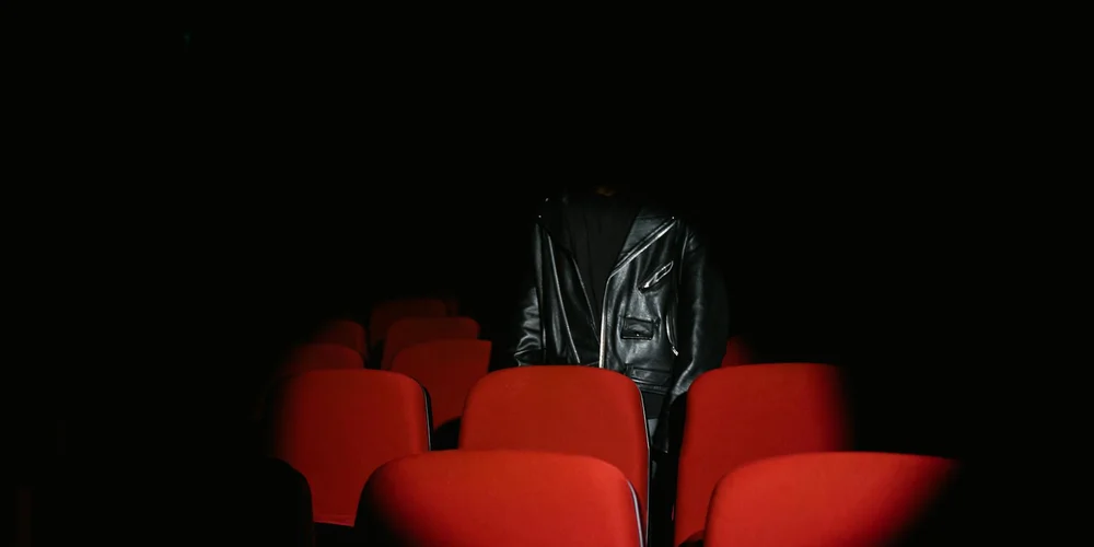 Person wearing a black leather jacket standing in a dim theater with red seats.