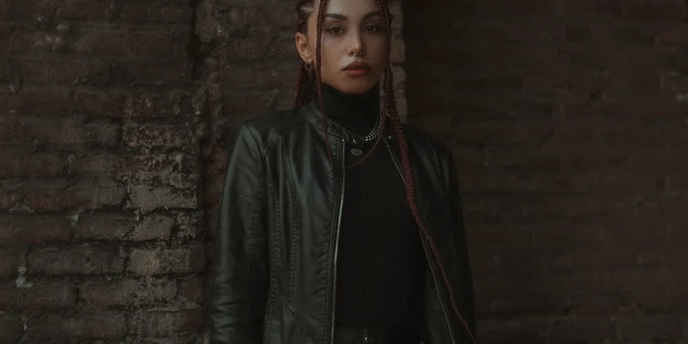 Person wearing a dark leather or faux leather jacket standing against a brick wall.