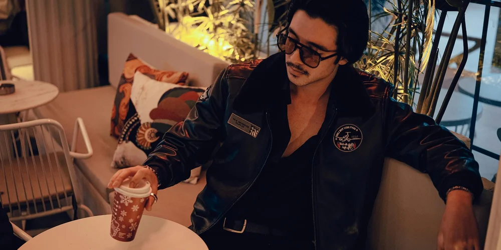 Man wearing a black leather jacket and sunglasses, seated in a cozy cafe with a coffee cup, presenting a smart-casual leather jacket ensemble.
