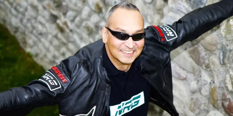 Person wearing a black leather jacket and sunglasses, arms outstretched, smiling outdoors