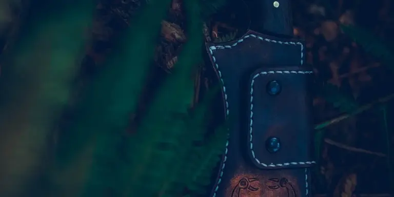 Close-up of a dark brown leather knife sheath with white stitching and two snaps, resting among green foliage