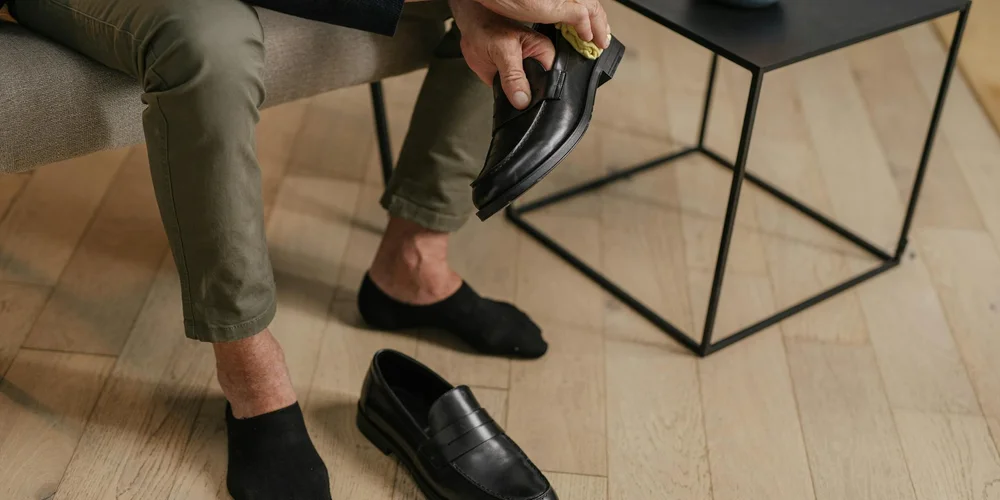 A person seated on a light wooden floor wearing olive-green pants and black socks, holding a black leather shoe while a small table sits nearby.