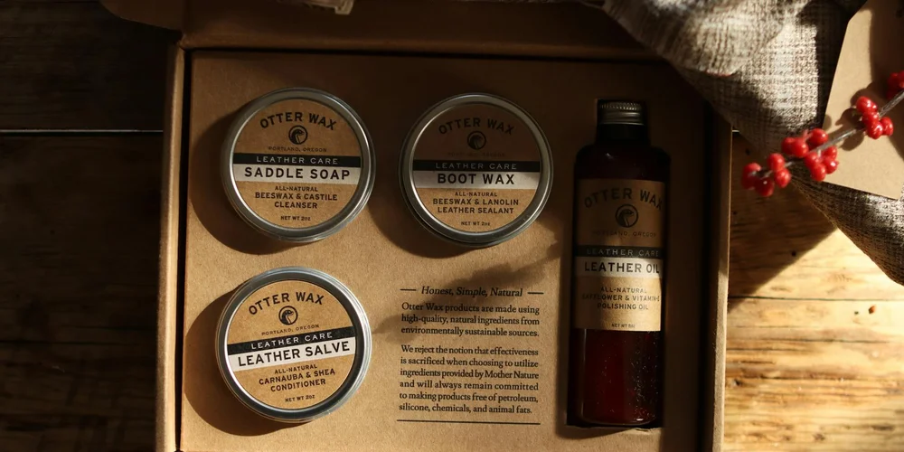 Assorted leather care products—two Otter Wax tins labeled Leather Salve and Saddle Soap, a bottle of leather oil, and other care items—on a wooden surface.