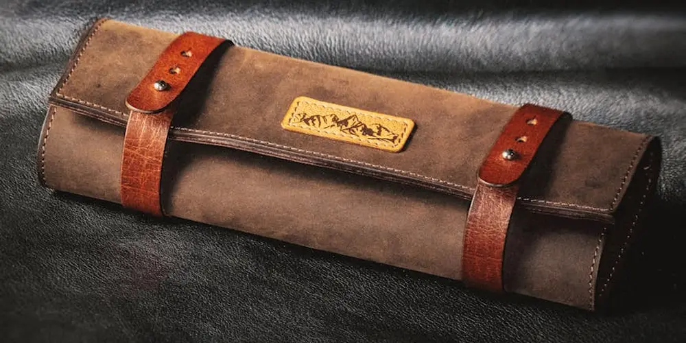 Brown leather roll-up case with two reddish-brown straps on a dark textured surface