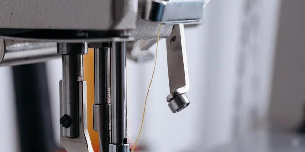 Close-up of a sewing machine needle and thread with metal components, suggesting leather stitching setup.