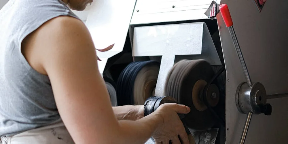 A person wearing a gray sleeveless shirt uses a bench-mounted polisher to buff leather on a workshop machine.