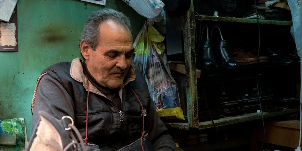 A man in a leather goods workshop stands beside shelves of leather shoes.