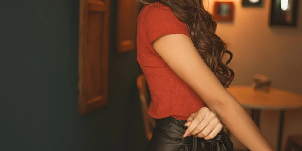 A person wearing a red short-sleeve top and a black leather skirt standing indoors.