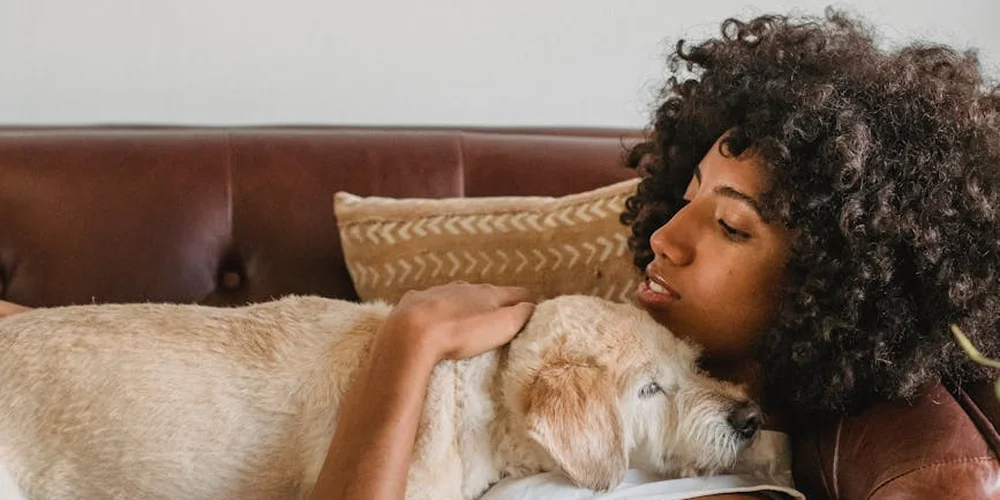 Person with curly hair cuddling a light-colored dog on a brown leather sofa