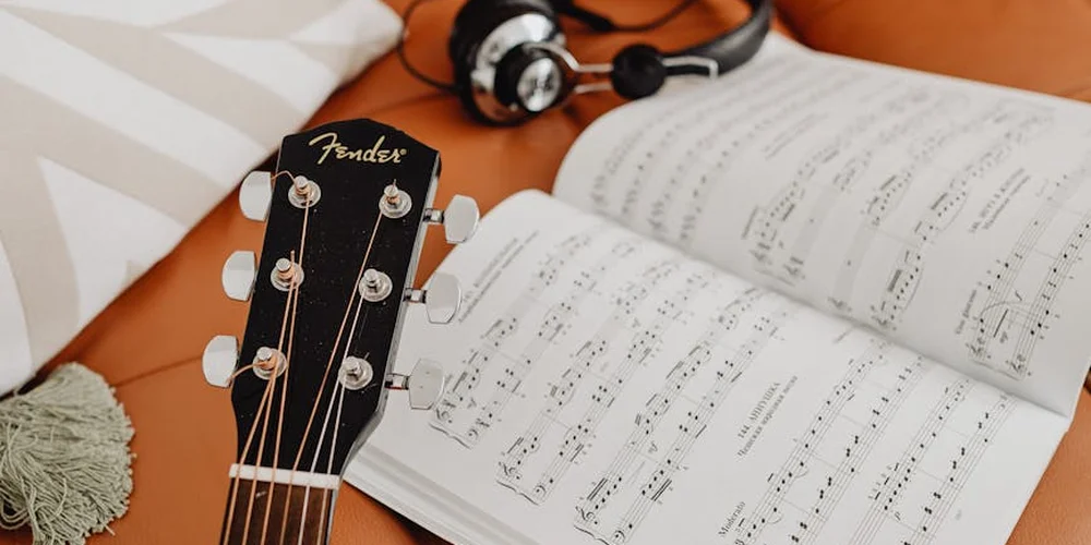 Close-up of a guitar headstock and headphones resting on an open sheet music book on a leather sofa, illustrating a calm post-repair care scene.