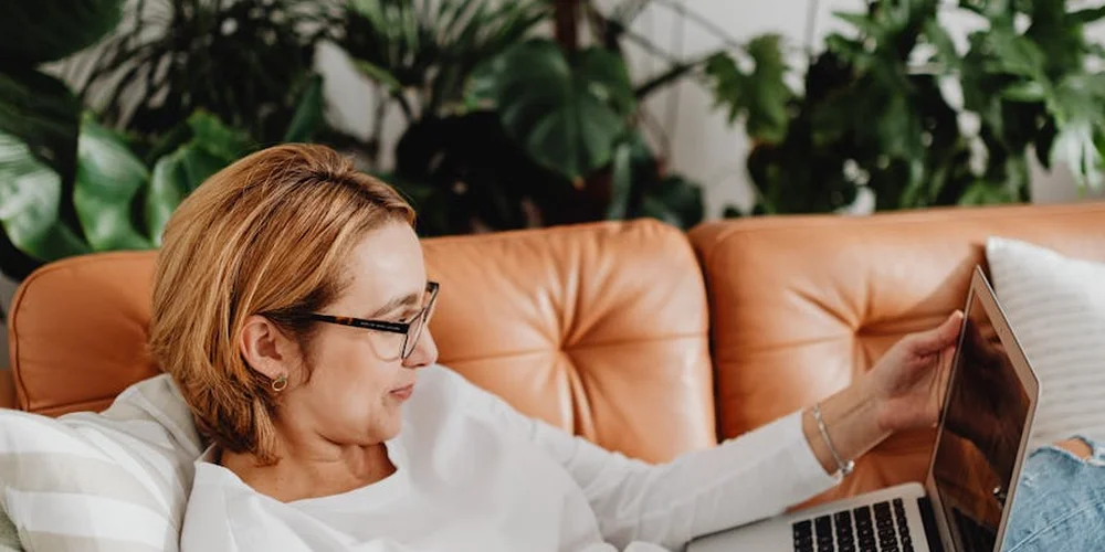 A woman with short blond hair and glasses sits on a tan leather couch, looking at a tablet.
