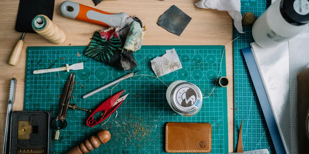 Leatherworking workspace with a green cutting mat, thread spools, scissors, pliers, and leather pieces prepared for stitching.