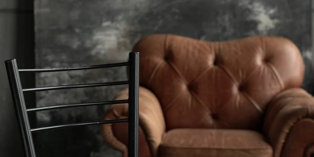 Dark, moody image of a brown tufted leather chair with a metal-framed chair in the foreground and a blurred dark background.