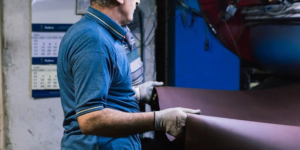 A leatherworker in a blue polo shirt and gloves handles a large sheet of leather in a workshop.