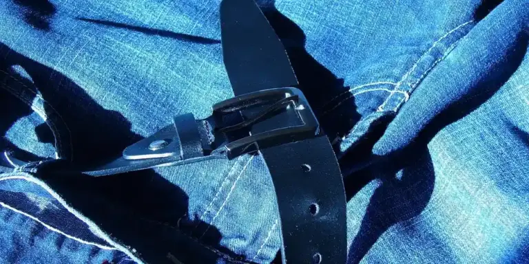Close-up of a black leather belt with multiple punched holes and a metal buckle resting on blue denim jeans.
