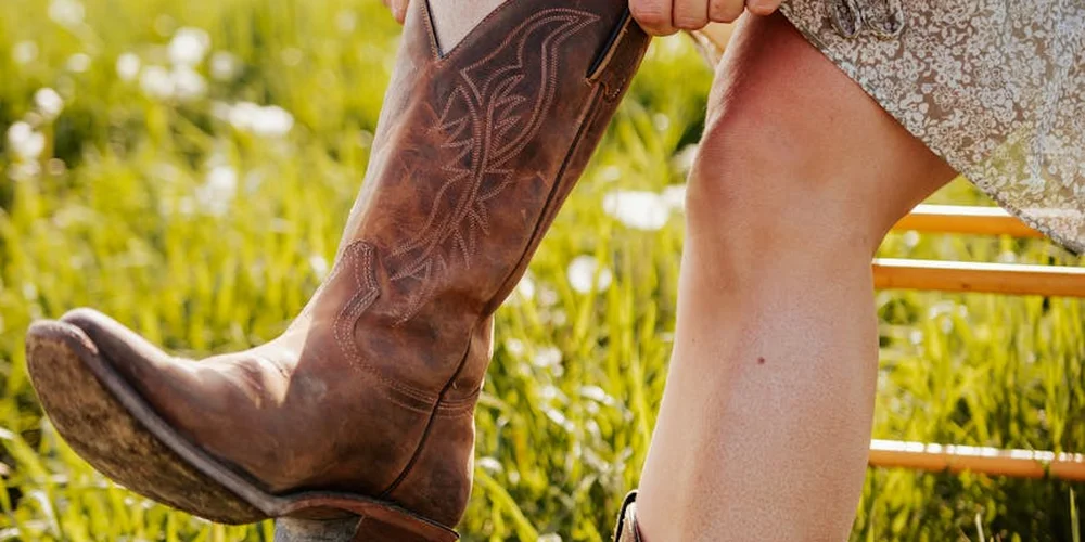 Close-up of a brown leather boot worn by a person outdoors, with a sunlit background and a bench in the distance.