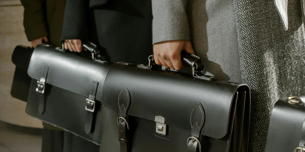 Hands gripping black leather briefcases, showcasing the durable, polished leather typical of chrome-tanned goods.