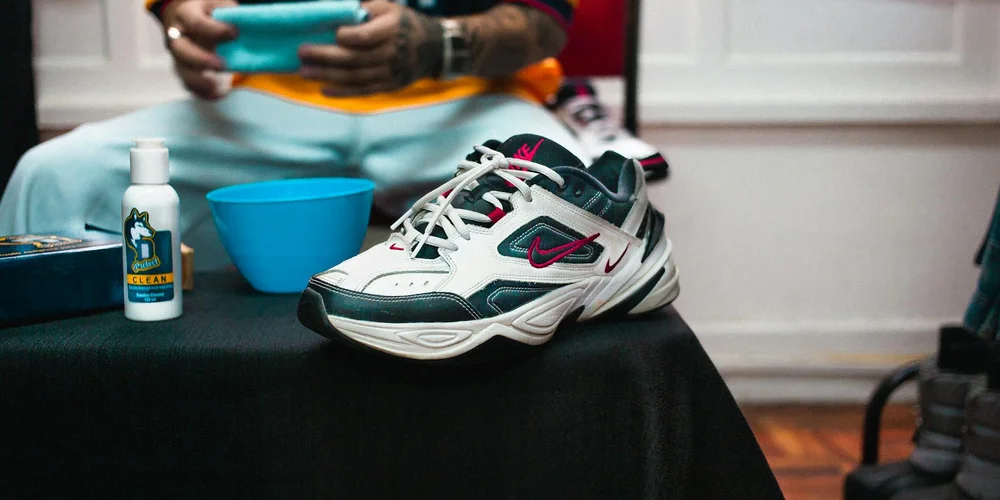 A sneaker on a black surface with cleaning supplies nearby and a person in the background preparing to clean.