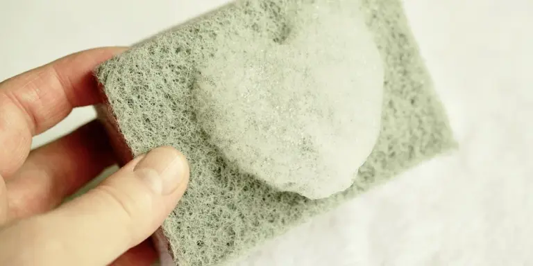 A hand holding a soft sponge against a light-colored leather surface, illustrating gentle cleaning.