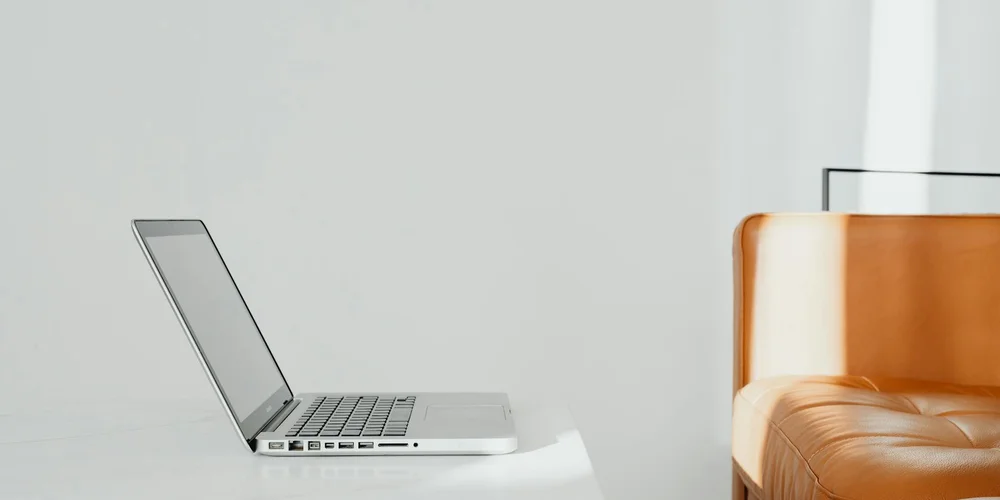 A laptop on a white desk beside a brown leather chair, illustrating the shift from traditional leather care to industrial-age products.
