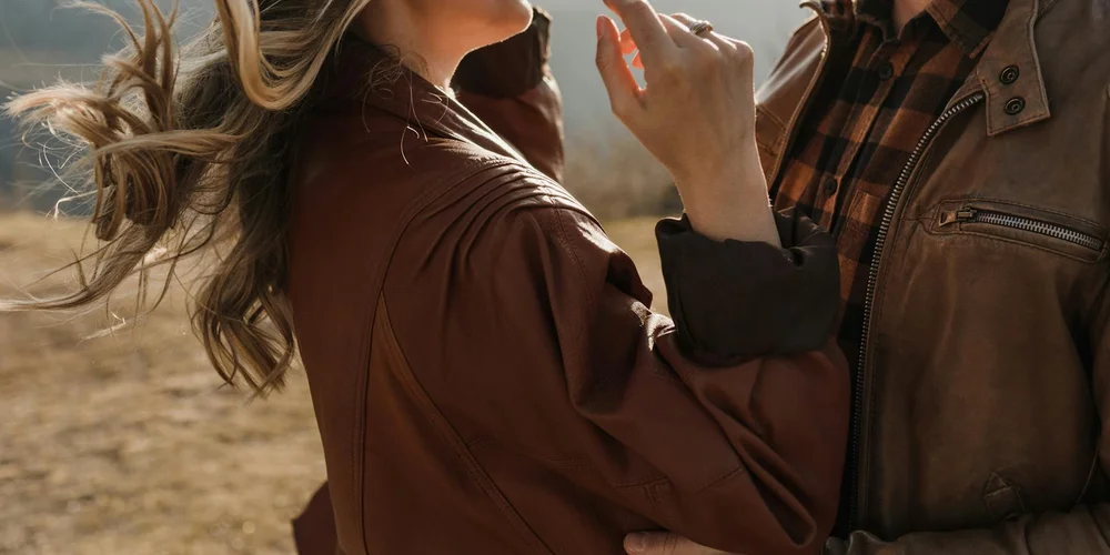 Two people wearing brown leather jackets outdoors; a close-up of hands and jackets, illustrating a hands-on approach to leather care.