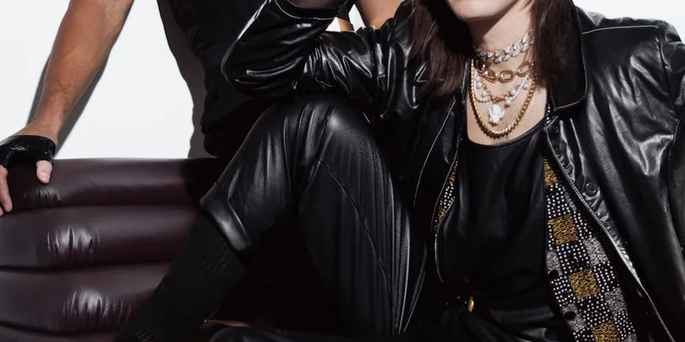 Two people in black leather outfits seated on a maroon leather couch, highlighting the texture and sheen of leather