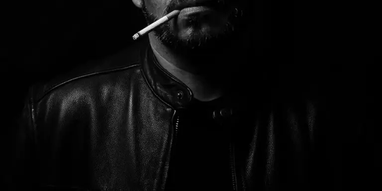 Close-up black-and-white photograph of a person wearing a black leather jacket with a cigarette in their mouth, highlighting the glossy leather texture and edgy mood.