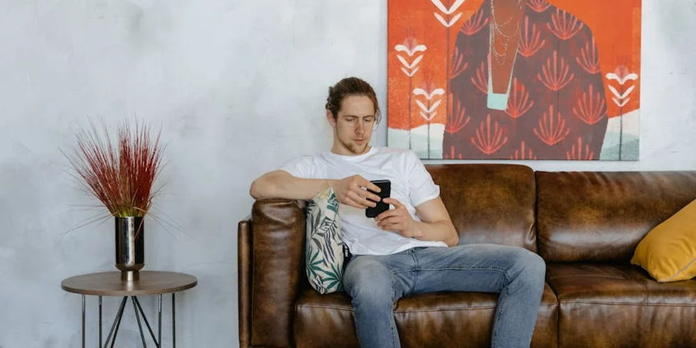 A man sits on a brown leather sofa, looking at his smartphone in a living room with a side table and modern wall art.