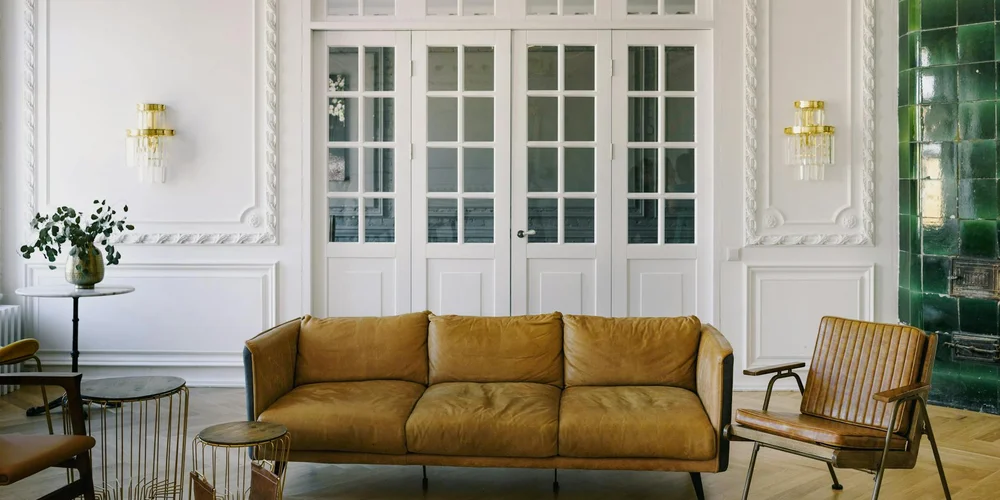 Tan leather sofa with matching chairs in a bright living room with white paneling and gold wall sconces.